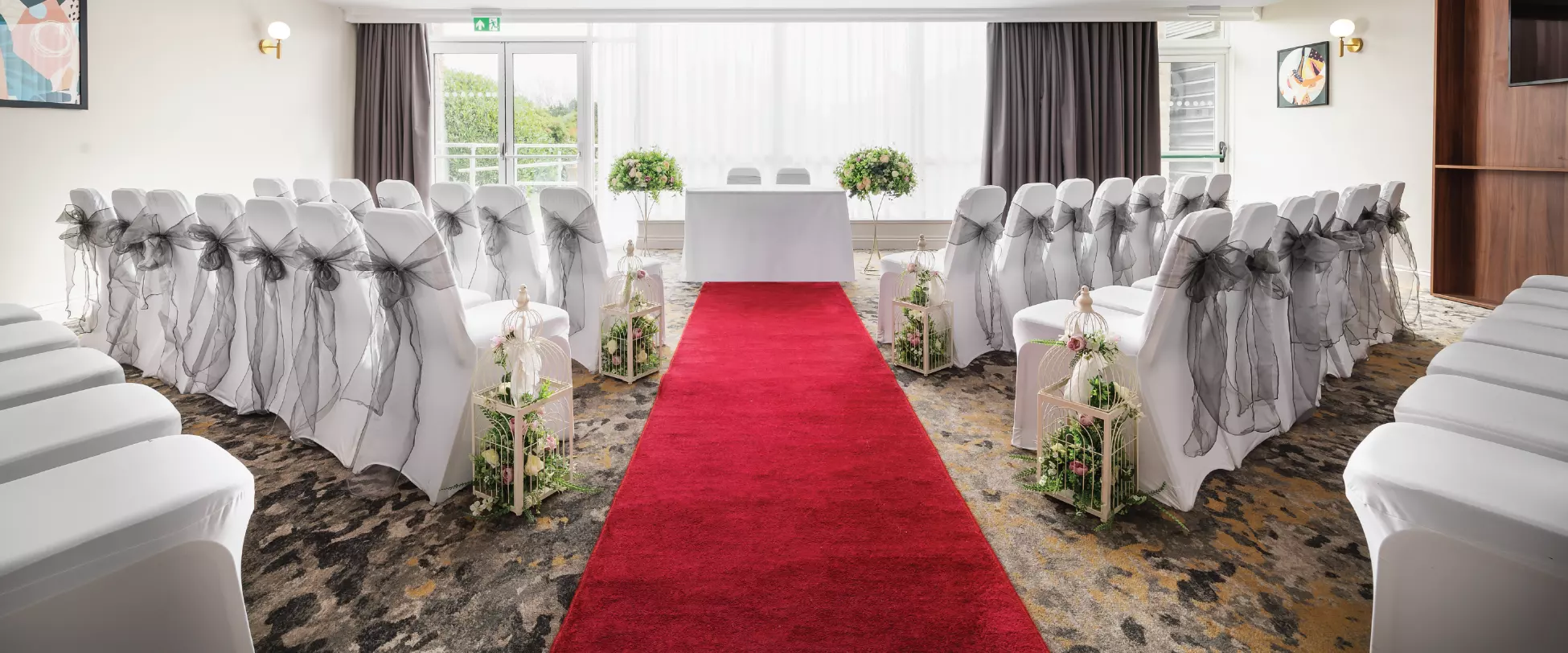 A wedding isle with a red carpet running up the centre of the room. Chairs with ribbons are on either side and there's a table in front of a big window..
